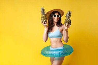 young curly slender brunette girl in a blue swimsuit in summer with an inflatable swimming circle holds pineapple in her hands and smiles, a woman with tropical fruits on vacation
