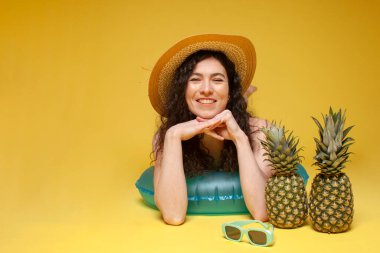 young girl in a straw hat lies on an inflatable swimming ring with pineapples and smiles on a yellow background, a woman sunbathes on vacation in the summer