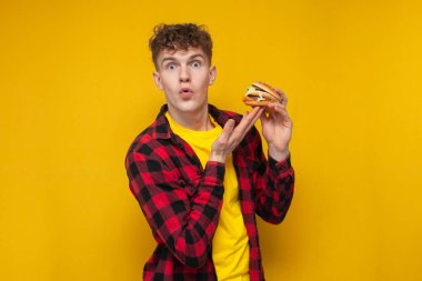 young curly guy student holding a burger on a yellow background and being surprised, man advertises fast food