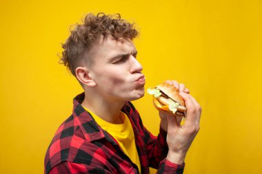 young curly guy student eats a burger on a yellow background and shows surprise, happy man with fast food