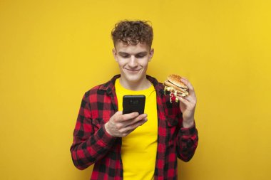 young curly guy holds a big burger and uses a smartphone, a student eats fast food and looks into the phone on a yellow background