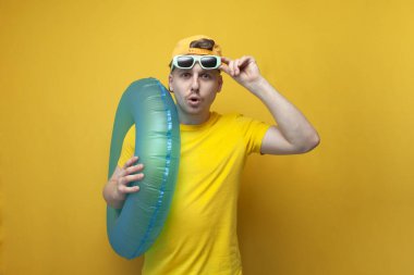 young surprised guy tourist in summer clothes and glasses holds a swim ring and wonders, vacation travel concept