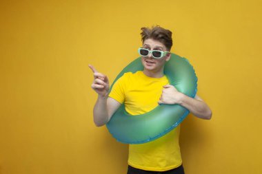young guy with a swimming inflatable ring points his finger to the side on a yellow background, a man tourist on vacation advertises mine space