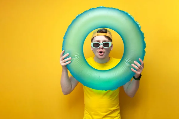 young surprised guy in glasses and a yellow t-shirt on vacation holds a swim ring and shows surprise, a man on a summer vacation, the concept of traveling to the sea