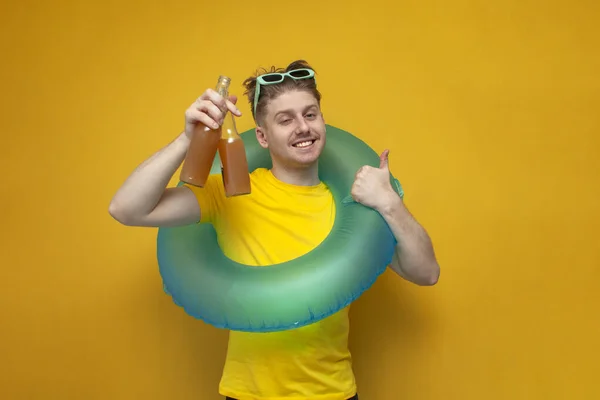 young happy guy in the summer on vacation shows bottles of beer and offers a drink on a yellow background, a man with an inflatable swim ring is resting on the sea with alcoholic drinks and shows like