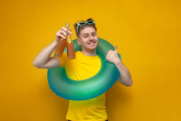 Молодой счастливый парень летом на отдыхе показывает бутылки пива и предлагает напиток на желтом фоне, человек с надувным плавательным кольцом отдыхает на море с алкогольными напитками и показывает, как