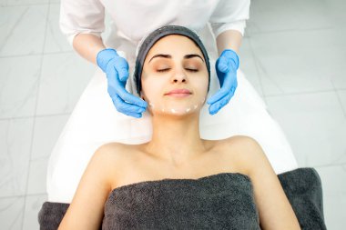 professional skin care in cosmetology clinic, dermatologist doctor applies cream on patient's face in clinic, cosmetologist makes face and head massage to girl