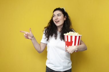 young curly girl viewer in white t-shirt holds popcorn and shows her hand to the side on yellow background, woman movie viewer advertises copy space