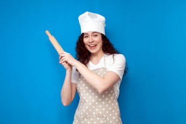 young girl chef in uniform holds kitchen item on blue background, woman housewife in an apron and hat on colored background