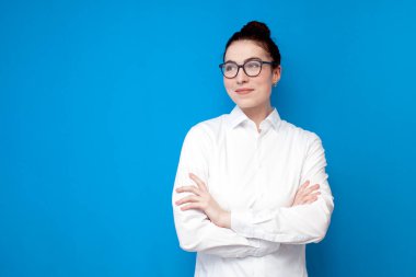 young girl office worker in white shirt and glasses stands with her arms crossed on blue background, female secretary and accountant on colored isolated background looks away at copy space