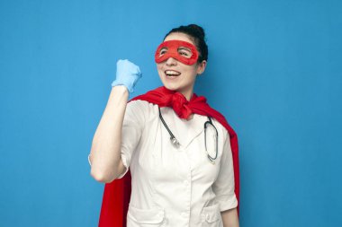 Süpermen kostümlü neşeli genç kız doktor mavi arka planda zaferle seviniyor, süper kahraman maskeli kadın hemşire kazanıyor.
