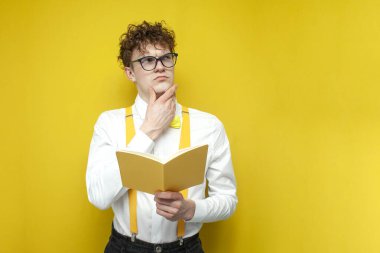 pensive nerd guy in glasses holds book and thinks on yellow isolated background, student in festive outfit reads book and looks up at copy space