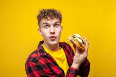 Genç kıvırcık erkek öğrenci sarı arka planda büyük lezzetli hamburgerler yer ve sürpriz, doymuş aç adam fast food yer.