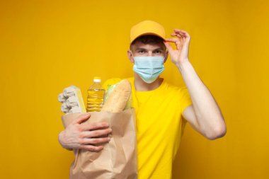 Sarı üniformalı ve tıbbi maskeli genç kurye sarı arka planda bir paket erzak tutuyor. Coronavirus salgınında yemek dağıtan adam.