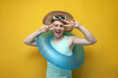 young guy in the summer on vacation with sunglasses and a swimming inflatable ring poses for the camera and shows a gesture of peace with his fingers on a yellow background