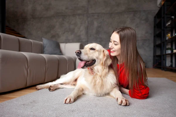 young girl plays with dog breed golden retriever at home on the floor, woman with pet together lies on mat near the sofa