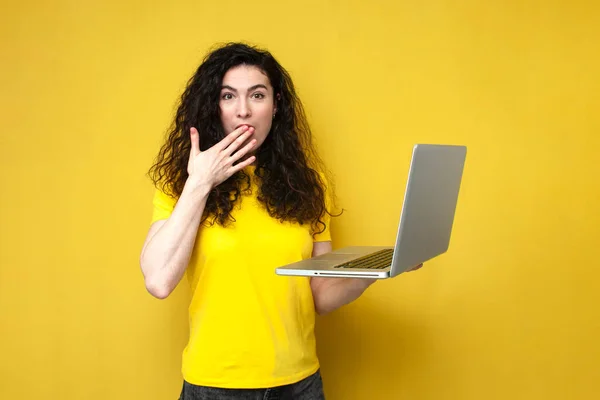 shocked curly girl in yellow t-shirt holding laptop and showing surprise covering her mouth with her hand on yellow background