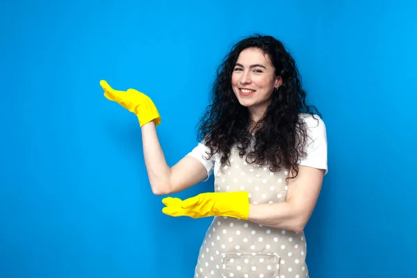 young woman cleaner in apron and gloves for cleaning shows her hands to the side on blue background, girl housewife advertises copy space, cleaning service worker on colored background