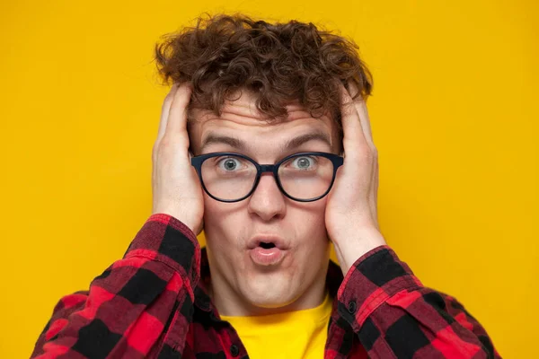 young curly shocked guy in glasses looks at the camera and holds his hands on his head on a yellow background, emotion of surprise, close-up