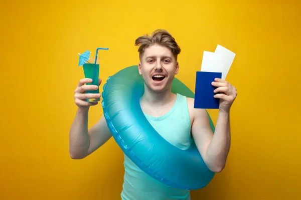 young happy guy with an inflatable swim ring holds a passport with tickets and a cocktail on a yellow background, a man flies for a vacation at sea concept