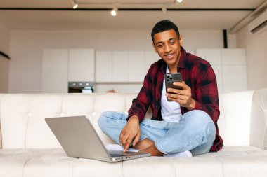 Genç Afrikalı Amerikalı adam evinde kanepede oturuyor ve dizüstü bilgisayar ve akıllı telefon kullanıyor. Erkek serbest çalışan odadaki telefona bakıyor, telefonu tutuyor ve bilgisayardan yazı yazıyor.