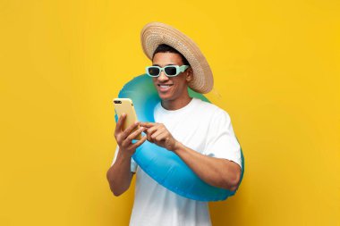 Şişme yüzme yüzüklü neşeli Afro-Amerikan genç adam sarı izole arka planda akıllı telefon kullanıyor. Yazın kumsalda yazan adam telefonla, seyahat ve yaz tatillerinde daktilo kullanıyor.