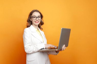 Gözlüklü ve beyaz takım elbiseli genç neşeli iş kızı renkli izole arka planda dizüstü bilgisayar kullanıyor, spor ceketli kadın yönetici bilgisayarda çalışıyor ve gülümsüyor.