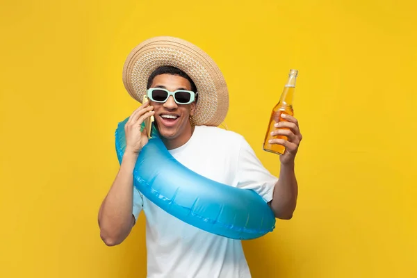 young guy afro american with inflatable swim ring holds bottle of beer and talks on the phone on yellow background, man in the summer on the beach communicates, concept of travel and summer holidays