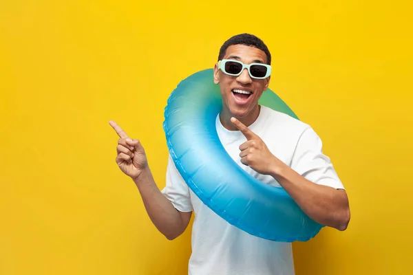african american guy in sunglasses with inflatable swim ring shows his hands to side on yellow isolated background, man in summer on beach advertises copy space, travel and summer vacation concept