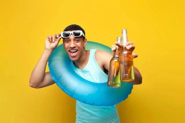 cheerful african american guy with inflatable swim ring holds two bottles of beer on yellow background, male tourist in summer at sea offers alcohol, concept of travel and summer vacation