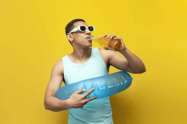 cheerful african american guy in sunglasses with inflatable swim ring drinks beer on yellow background, male tourist in the summer at sea with alcohol, concept of travel and summer vacation