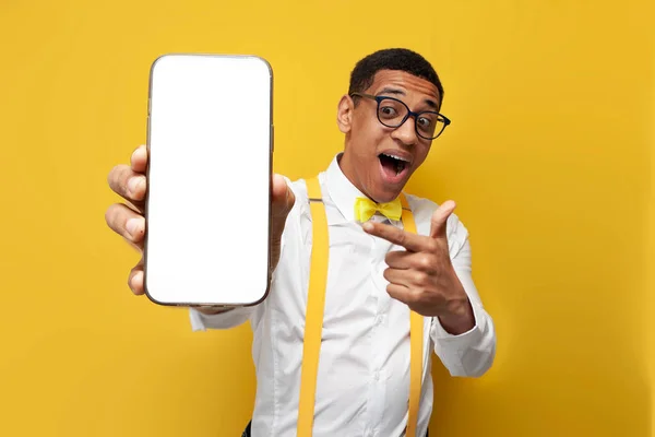 young afro american guy in festive outfit with bow tie and suspenders shows blank screen of smartphone on yellow background, nerd man advertises phone, mock-up with smartphone screen