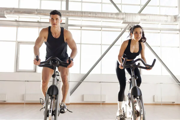 beautiful athletic couple of cyclists train in the fitness room on static bicycle simulator, man trainer and fitness woman go in for sports together on simulators in the morning in bright room
