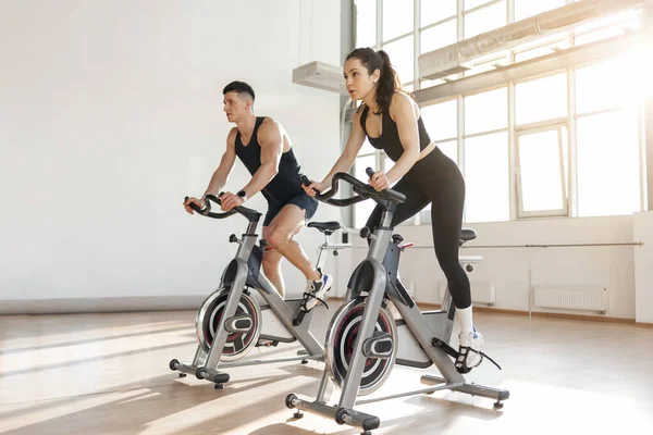 beautiful athletic couple of cyclists train in the fitness room on static bicycle simulator, man trainer and fitness woman go in for sports together on simulators in the morning in bright room