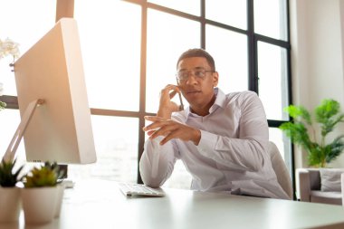 Gömlekli Afro-Amerikalı bir adam işyerindeki ofisinde oturuyor ve telefonda konuşuyor. İş elbiseli bir işadamı elini monitöre gösteriyor ve akıllı telefonuyla iletişim kuruyor.
