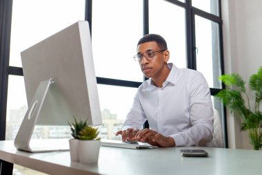 Gözlüklü ve gömlekli Afrikalı bir ofis çalışanı masaya oturur ve bilgisayarda yazar, işadamı internette çalışır ve monitöre, işyeri yöneticisine bakar.