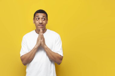 Beyaz tişörtlü Afro-Amerikan genç adam çok şey umuyor ve soruyor sarı izole arka planda katlanmış ellerle, adam yalvarıyor, bekliyor ve umut veriyor ve yalvarıyor.
