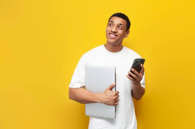 Beyaz tişörtlü genç Afro-Amerikalı adam elinde dizüstü bilgisayar tutuyor ve akıllı telefon kullanıyor ve sarı izole bir arka plan hayal ediyor modern aletlerle fotokopi alanına bakan dalgın bir adam.