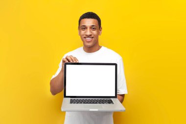 Beyaz tişörtlü genç Afro-Amerikalı adam sarı izole edilmiş arka planda boş dizüstü bilgisayar ekranı gösteriyor.