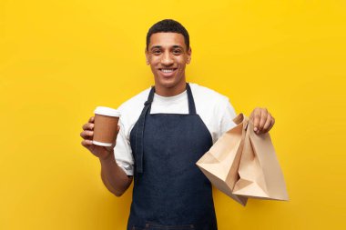 Üniformalı genç Afrikalı Amerikalı barista sarı arka planda kahve ve paket yiyecek tutuyor.