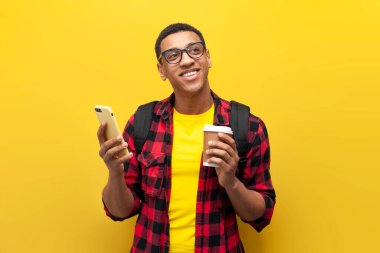 Sırt çantalı Afrika kökenli Amerikalı genç bir öğrenci kahve tutuyor ve sarı izole edilmiş arka planda akıllı telefon kullanıyor, cep telefonuyla hayal kuran Latin bir öğrenci, öğrenim ve eğitim kavramı
