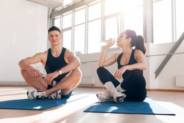 young athletic couple in sportswear in training sit on yoga mat and drink water from bottle, man and woman do fitness in the morning and maintain water balance