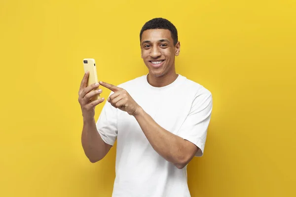 stock image young african american man in white t-shirt uses smartphone and presses his finger on the screen on yellow isolated background, man chooses online shopping by phone