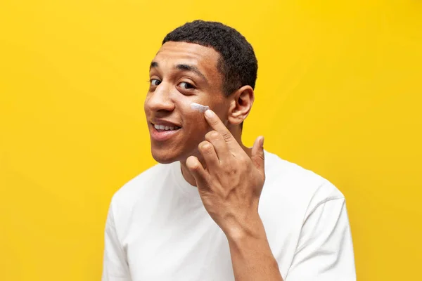 stock image young african american guy in white t-shirt applies cream on his face on yellow isolated background, man uses facial cosmetics and takes care of his skin