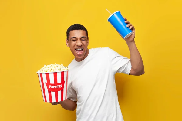 stock image young happy guy african american rejoices and dances with popcorn and soda on yellow isolated background, satisfied male cinema viewer smiles