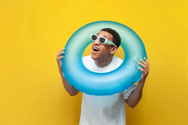 shocked african american guy in sunglasses with inflatable swim ring is surprised on yellow background, male tourist in the summer looks away at copy space, concept of travel and summer vacation