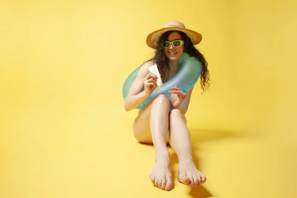 stock image young girl in a blue swimsuit with a swimming inflatable ring sits on the beach and holds sunscreen on a yellow background, a woman on vacation uses sunscreen in the summer