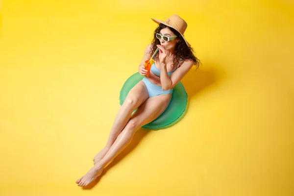 stock image young girl in blue swimsuit sits on inflatable swimming ring holds orange alcoholic cocktail on yellow background, woman sunbathes on vacation in the summer