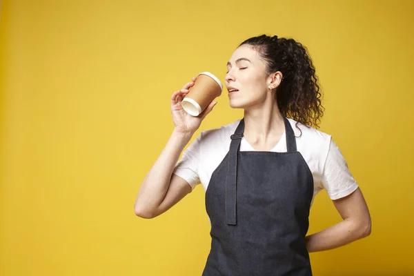 デニムのエプロンの少女は黄色の背景にコーヒーを飲む制服を着た女性のウェイターの肖像画 コピースペース — ストック写真