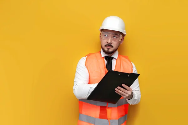 asian civil engineer in uniform writes plan on paper and looks away on yellow isolated background, thoughtful male korean man in hardhat and work vest meditates, worker architect has an idea